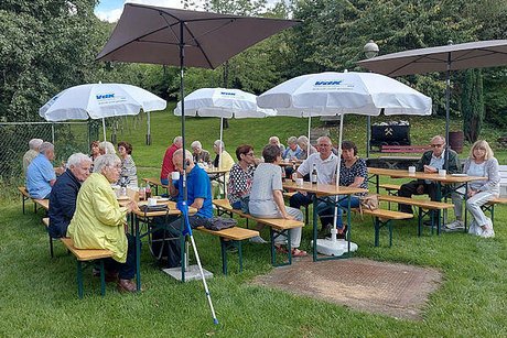 Eine Gruppe Männer und Frauen sitzt an Bierzeltgarnituren, die auf einer großen Wiese aufgebaut sind, unter zwei eckigen und drei weißen Sonnenschirmen mit VdK-Logo. Im HIntergrund steht eine alte Bergwerkslore in der Wiese.