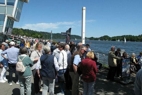 Viele Menschen stehen dicht beieinander am Ufer des Baldeneysees und warten auf das Ausflugsschiff. Links im Bild der verglaste Turm der Regattabahn.