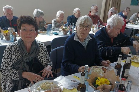 Zwei Frauen und ein Mann sitzen beim Essen an einem Tisch nebeneinander. Im Hintergrund sitzen weitere Menschen an langen Tischen beim Essen.