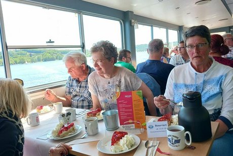 Menschen sitzen bei Getränken und Kuchen an Tischen des Ausflugschiffes.