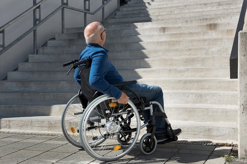 Ein &auml;lterer Mann im Rollstuhl steht ratlos am Fu&szlig; einer hohen Treppe, er schaut die Stufen hinauf. 
