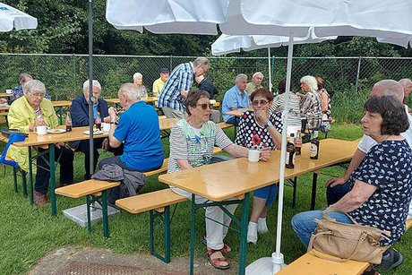 Eine Gruppe Menschen sitzt gemeinsam unter Schirmen an Bierzeltgarnituren, die auf einer Wiese aufgebaut sind.