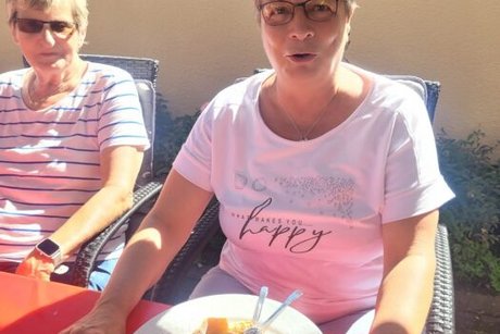 Zwei Frauen sitzen nebeneinander an einem Tisch. Die Frau rechts im Bild hat einen Teller mit einer üppig mit Früchten belegten Waffel vor sich stehen und macht ein staunendes Gesicht Richtung Kamera.