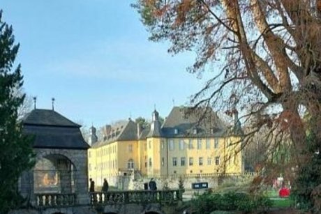 Im Vordergrund der Wassergraben, darüber führt eine Brücke zum Schloss, dass in der Wintersonne leuchtet. Rechts im Bild ein Baum mit orangerotem Herbstlaub.