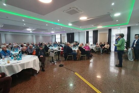 Viele Menschen sitzen an Tischen in einem Saal und werden von einem Mann mit Mikrofon in der Hand begrüßt.