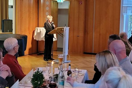 Viele Menschen sitzen gemeinsam an einem langen Tisch. Im Hintergrund eine Frau am Rednerpult.