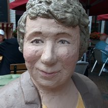 Die Skulptur Alltagsmensch in Straelen ist eine Frau mit grauem lockigen Haar, grauer Jacke und gelbem Shirt.