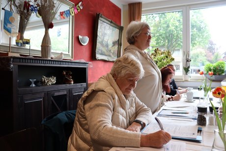 Eine Frau sitzt am Tisch und schreibt etwas, dahinter steht eine Frau und spricht zu anderen Menschen.