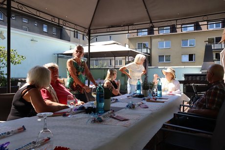 Mehrere Personen sitzen an einem Tisch und andere stehen daneben und unterhalten sich miteinander.