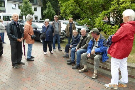 Eine Gruppe Männer und Frauen wartet auf einem Platz. Rechts im Bild sitzen drei der Männer auf einer Mauer, die ein Stück Wiese mit Busch einfasst.