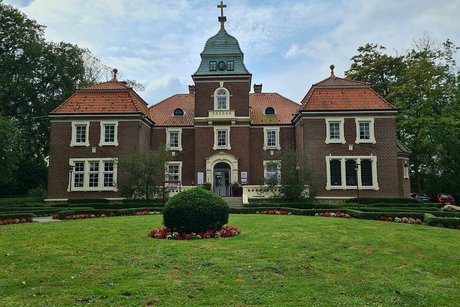 Ein roter, von zwei Seitenflügeln gesäumter Ziegelbau mit Turm in der Mitte. Im Vordergrund eine große Wiese mit einzelnem Buchsbaum.