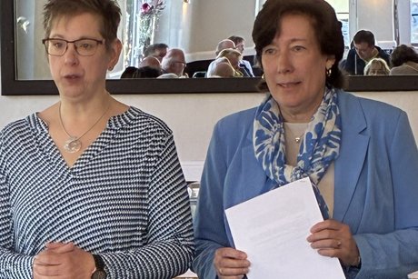 Vorsitzende und Schriftführerin beantworten Fragen zum Protokoll der letzten Versammlung zwei Frauen stehen in einem Saal