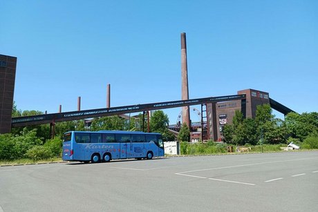 Ein blauer Reisebus steht auf einem ansonsten menschen- und fahrzeugleeren Parkplatz. Im Hintergrund sind einige rote Ziegeltürme und Förderbandanlagen zu erkennen.