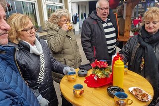 Menschen stehen um einen Stehtisch aus Holz herum und essen Wurst und trinken Heißgetränke aus Tassen.