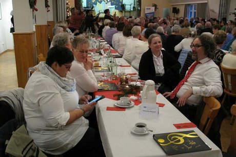Viele Menschen sitzen in einem Saal an langen, weihnachtlich geschmückten Tischen.
