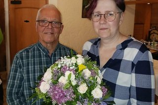 Ein Mann und eine Frau stehen nebeneinander. Die Frau hält einen großen Strauß mit weißen und lila Blumen.