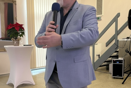 Ein Mann mit hellgrauem Sakko, Bart und Brille steht und spricht in ein Mikrofon, das er in den H&auml;nden h&auml;lt.