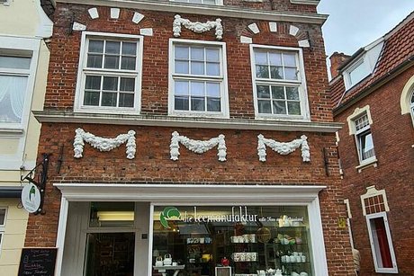 Mit weißem Stuck verzierte Außenfassade eines alten Giebelhäuschens in der Auricher Innenstadt. Im Erdgeschoss befindet sich ein Ladengeschäft mit großem Schaufenster.