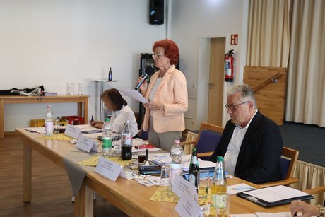 Jahreshauptversammlung 2026 Eine Frauen und ein Mann sitzen an einem Tisch, dazwischen steht eine Frau und liest einen Bericht vor.