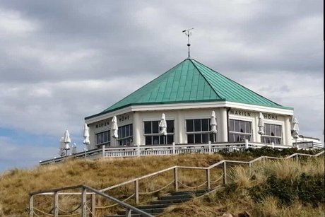 Über eine Treppe durch die Dünen erreichbar, liegt ein weißer Strandpavillon mit grünem Kupferdach.
