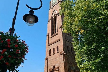 Der Kirchturm einer roten Backsteinkirche in Jever ragt neben einem großen grünbelaubtem Baum in den wolkenlosen Himmel. Links im Bild eine altmodische Straßenlaterne mit Blumenampel.
