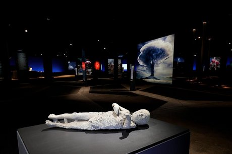Eine weiße menschliche Skulptur liegt auf einem schwarzen Podest, im Hintergrund Fotos einer Atombombenzündung.