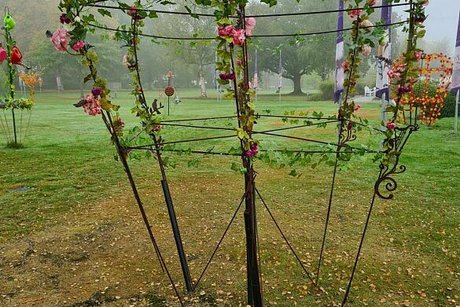 Schwarze Metallbügel sind in Kronenform gebogen in eine Wiese gesteckt und mit Blumen und Efeu geschmückt.