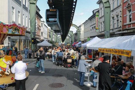 Links im Bild ein buntes Kinderkarussel. Rechts einige Pavillons und Stände, umringt von vielen Besuchern. Oben im Bild fährt gerade die Schwebebahn an der Schiene entlang.