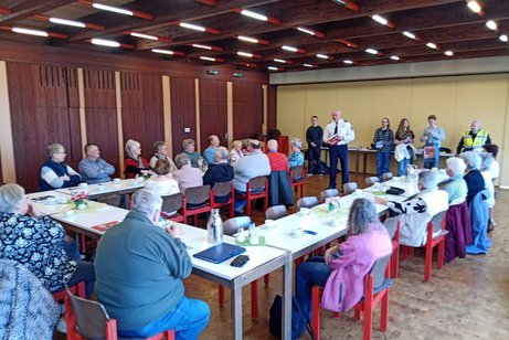 Viele Menschen sitzen an langen Tischen zusammen und h&ouml;ren einem Redner zu.