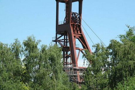 Das Wahrzeichen von Essen: der rostig-rote Förderturm von Zeche Zollverein hebt sich deutlich vor blauem Himmel ab. Im Vordergrund grüne Birken und Büsche.