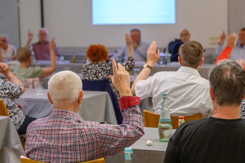 Menschen sitzen an Tischen in einem Saal und heben die Hand zur Abstimmung.