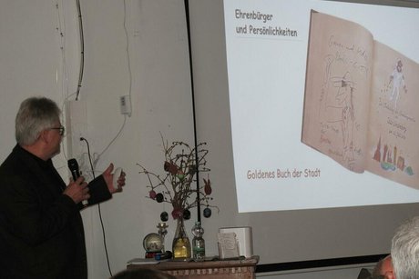 Ein Mann mit Mikrofon steht vor einer Leinwand, auf der eine Powerpoint-Präsentation gezeigt wird: aktuell das Foto eines aufgeschlagenen Buches.