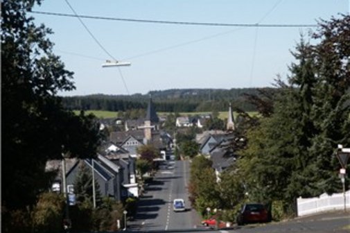 Blick auf die Ortschaft Wilnsdorf mit einer breiten, von Fichten gesäumten Straße, die hügelabwärts führt, den Dächern des Ortes und zwei Kirchtürmen.