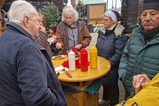 Menschen stehen um einen Stehtisch aus Holz herum und essen Wurst.