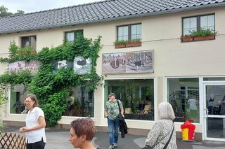 Menschen stehen im Innenhof der Straußenfarm vor hellgestrichenen Gebäuden an denen gemalte Bilder von Straußen hängen.