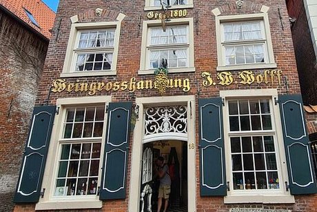 Außenfassade eines alten Giebelhauses aus roten Ziegeln mit weißen Sprossenfenstern und grünen Schlagläden. Über der Holztür prangt in goldenen, altdeutschen Lettern der Schriftzug Weingroßhandlung J.W. Wolff
