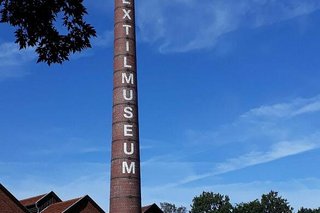 Ein großer Ziegelschornstein erhebt sich vor blauem Himmel mit weißer Aufschrift: Textilmuseum