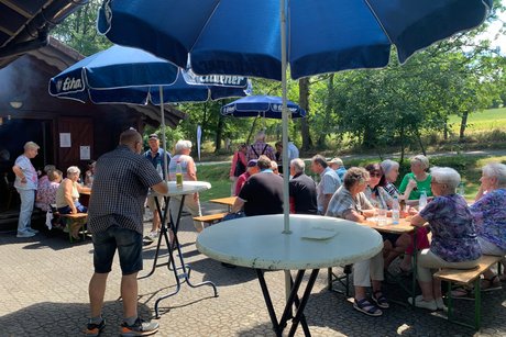 Personen an Tischen mit Sonnenschirmen im Gr&uuml;nen vor einer Grillh&uuml;tte