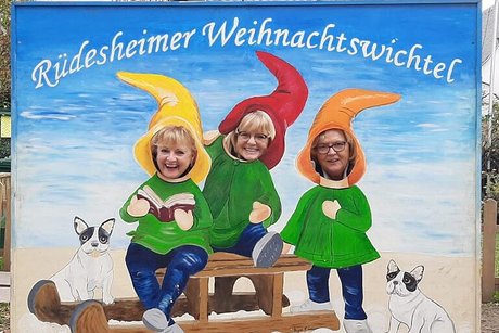 Drei Frauen posieren hinter einer Stellwand mit Gucklöchern, auf der in bunten Farben drei Rüdesheimer Weihnachtswichtel bei der Schlittenfahrt aufgemalt sind.