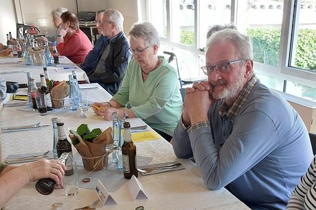 Menschen sitzen an einem langen Tisch nebeneinander. Auf dem Tisch stehen verschiedene Getränkeflaschen. Hinter ihnen erkennt man eine Reihe Fenster.