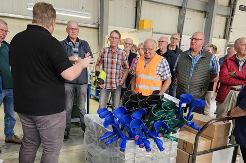 Mitarbeiter steht vor einer Gitterbox mit Zubehör für Gehstützen und erklärt 20 Teilnehmern des OV Dreierwalde die Herstellung von Zubehör für die Gehstützen
