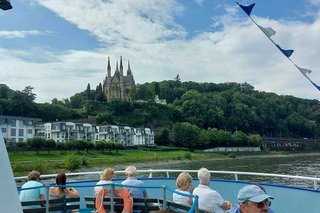 Menschen sitzen auf dem Außendeck eines Ausflugsschiffes. Am linken Ufer stehen Neubauhäuser. Dahinter erhebt sich zwischen Bäumen die Kirche St. Apollinaris.