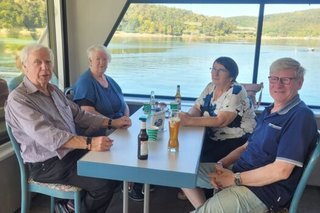 Zwei Frauen und zwei Männer sitzen gemeinsam an einem Tisch an Deck des Ausflugsschiffs. Durch das Fenster hinter ihnen erkennt man den Edersee und das umliegende, bewaldete Ufer.