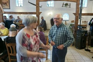 Ein Mann und eine Frau tanzen gemeinsam auf einer freien Fläche des Veranstaltungsraumes. Links im Hintergrund sitzen Menschen an Tischen.