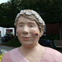 Das Bild zeigt eine Skulptur, den Oberkörper einer Frau mit grauem Haar und rosa Kleid.