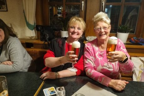 Zwei Frauen sitzen an einem Tisch und prosten lächelnd mit zwei Gläsern Bier in die Kamera.