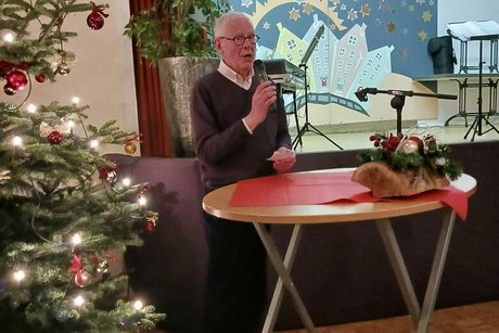 Ein Mann mit Mikrofon steht an einem weihnachtlich geschmückten Stehtisch. Hinter ihm eine leere Bühne, links von ihm steht ein geschmückter Weihnachtsbaum.