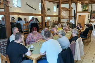 Menschen sitzen in einer Gaststube an einem langen Tisch.