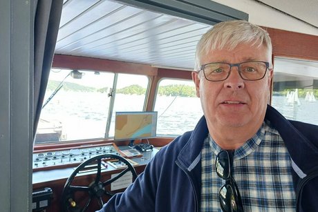 Portrait eines weißhaarigen Mannes mit Hemd und Brille. Im Hintergrund das Steuerrad des Schiffes.