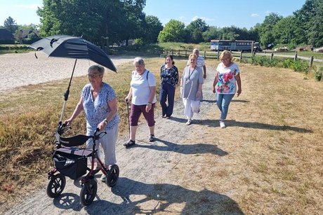 Eine Gruppe Menschen läuft einen sandigen Weg durch vertrocknete Graslandschaft entlang. Vorneweg eine Frau mit Rollator und Schirm, der vor der strahlenden Sonne schützt.